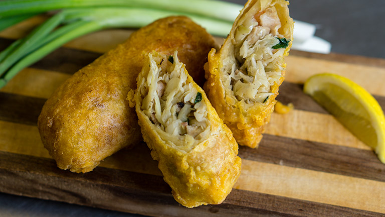 Egg rolls, one full and one cut in half, sitting on a striped cutting board with a lemon and green onions